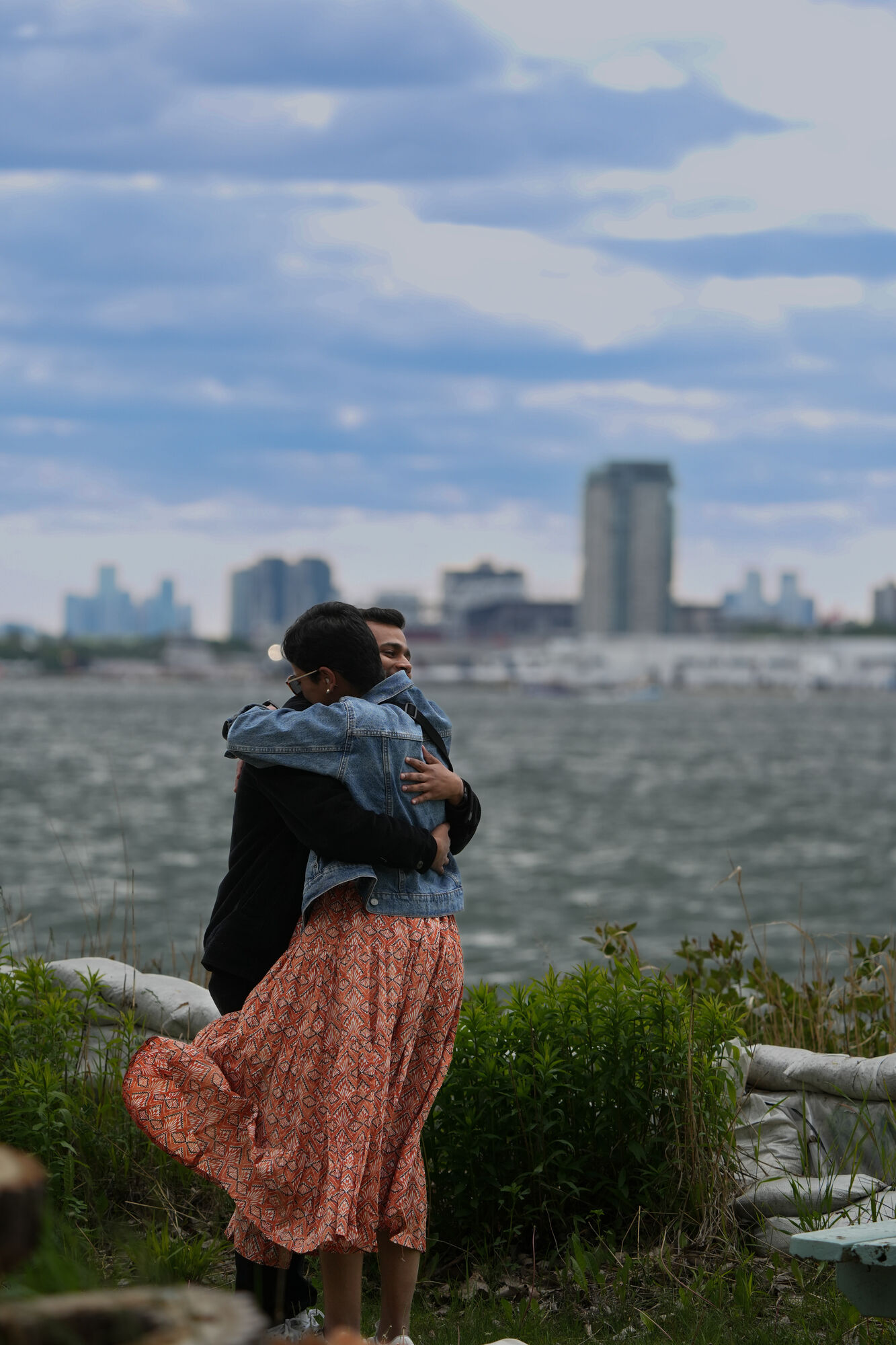 Soumen and Nikita embracing by the waterfront, city skyline in the background